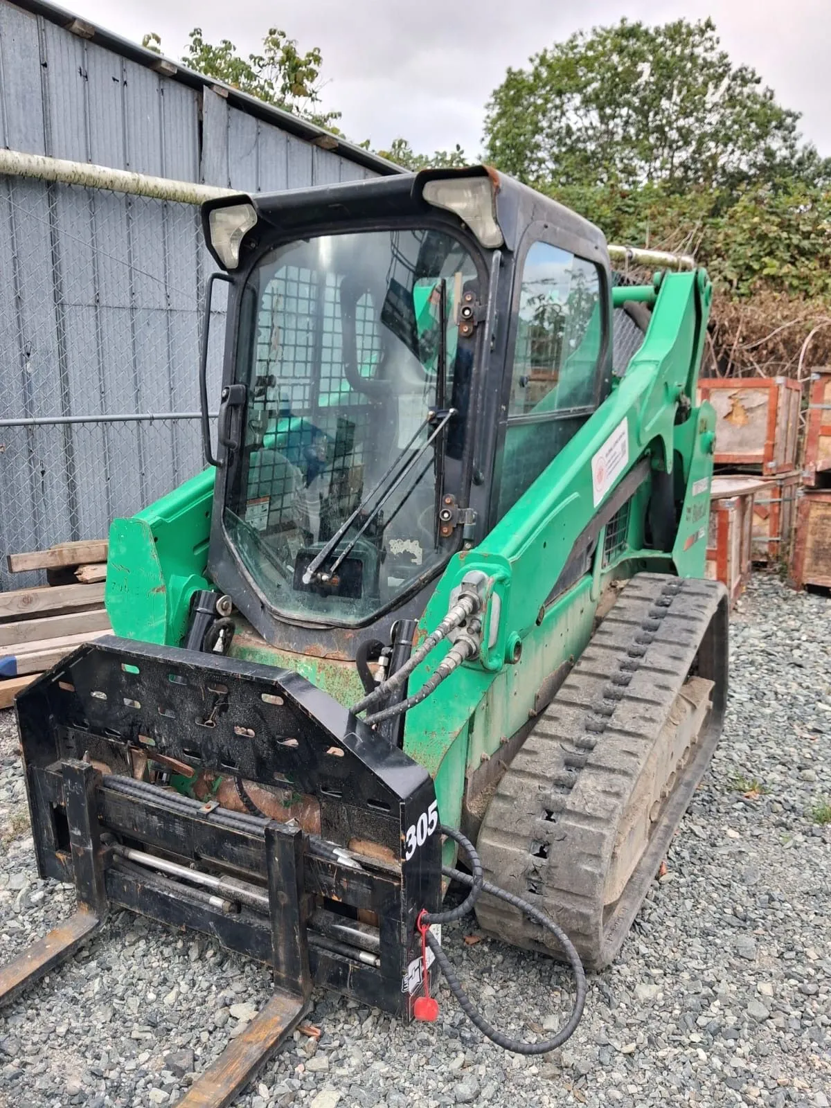 Green tracked skid steer with forks parked beside a yard building